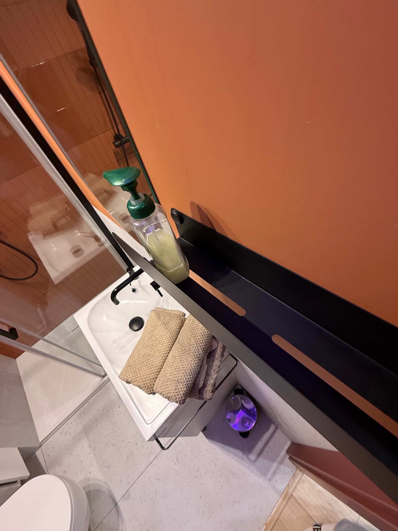 Umywalka w łazience z dozownikiem mydła, ręcznikami i czarną półką na tle pomarańczowej ściany. Widok z góry, oświetlenie punktowe.