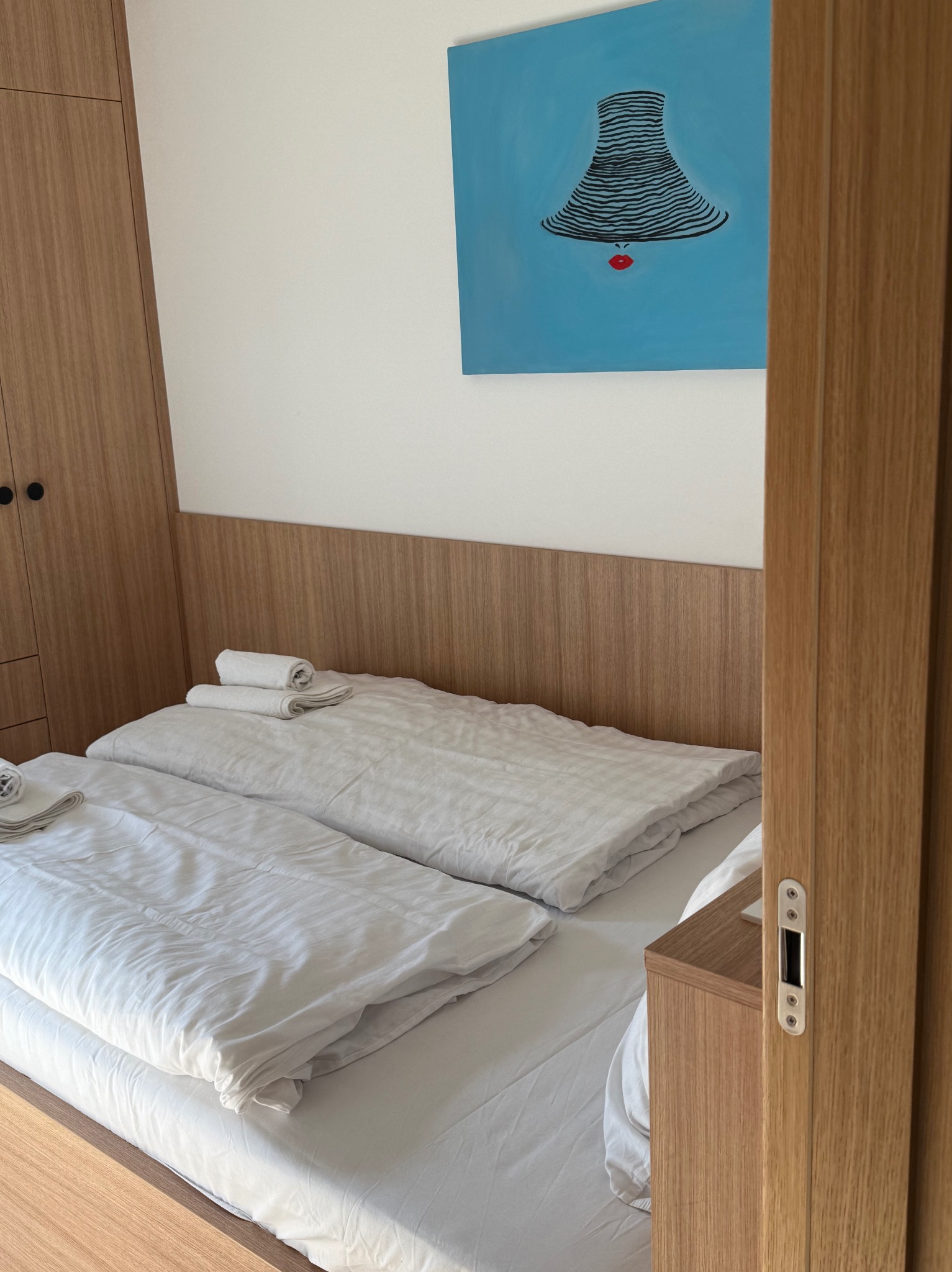 Czysta, biała pościel na dwóch łóżkach w pokoju hotelowym, z minimalistycznym obrazem na ścianie i drewnianymi meblami. Widok zza uchylonych drzwi.