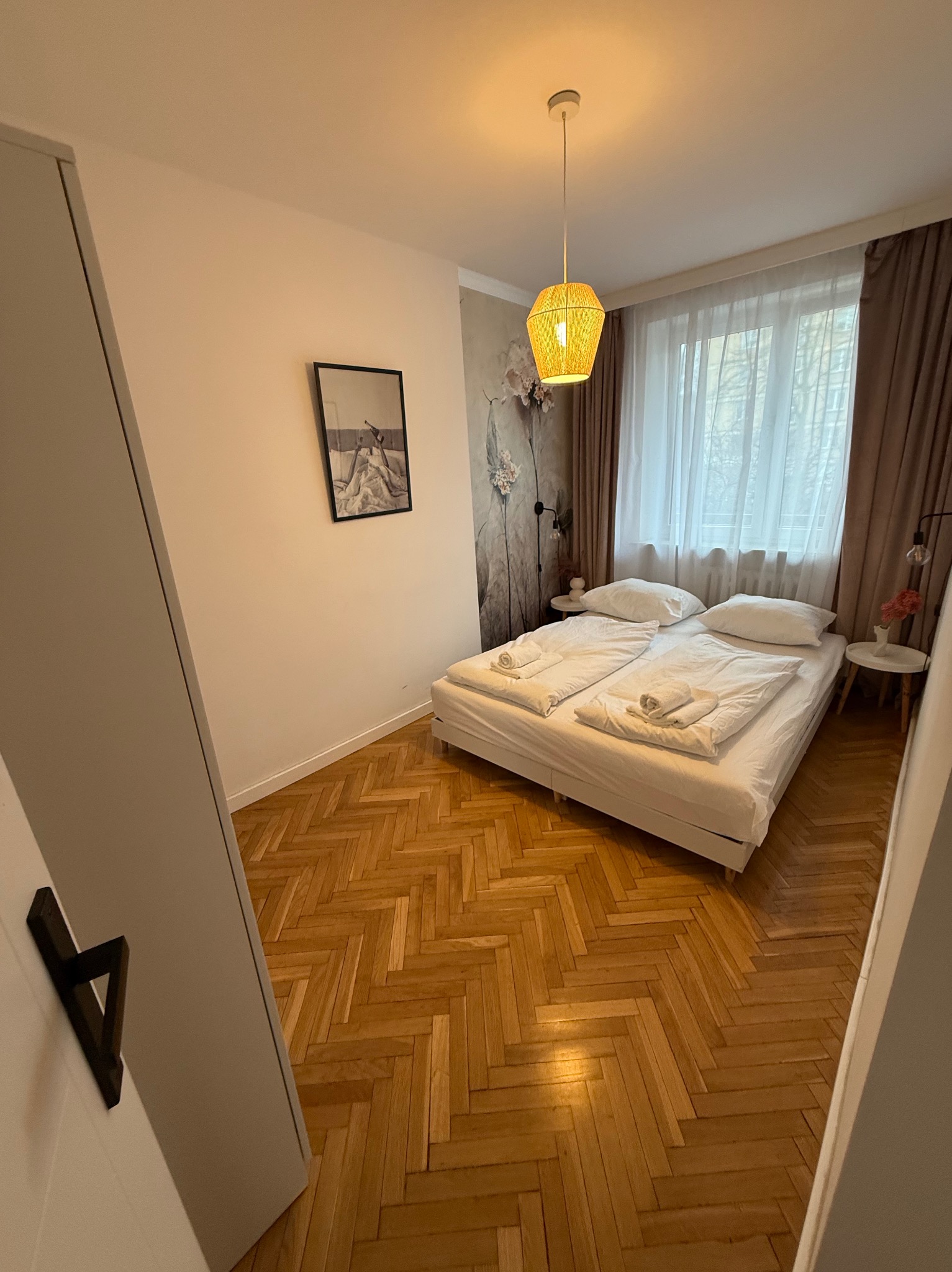 Jasna sypialnia z parkietem w jodełkę, białym łóżkiem z ręcznikami, lampą z abażurem i szafą. Na ścianie grafika i kwiaty. Widok zza uchylonych drzwi.