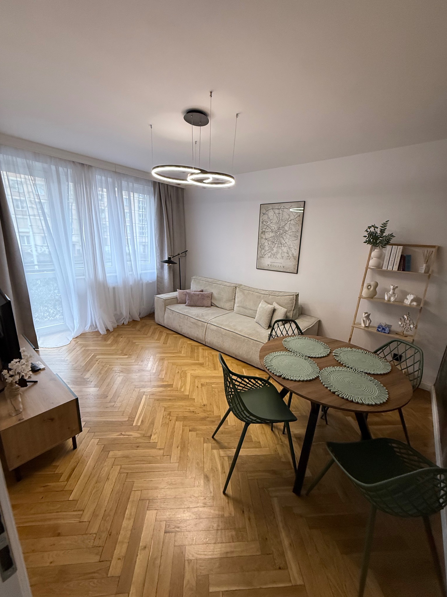 Jasne wnętrze mieszkania z parkietem w jodełkę, beżową sofą i okrągłym stołem z zielonymi podkładkami. Nowoczesna lampa sufitowa i plakat na ścianie.