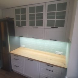 Dawid - Jasna kuchnia z drewnianym blatem i białymi szafkami z przeszklonymi frontami. Podświetlenie LED nad blatem. Nowoczesny design z ciemnymi uchwytami.