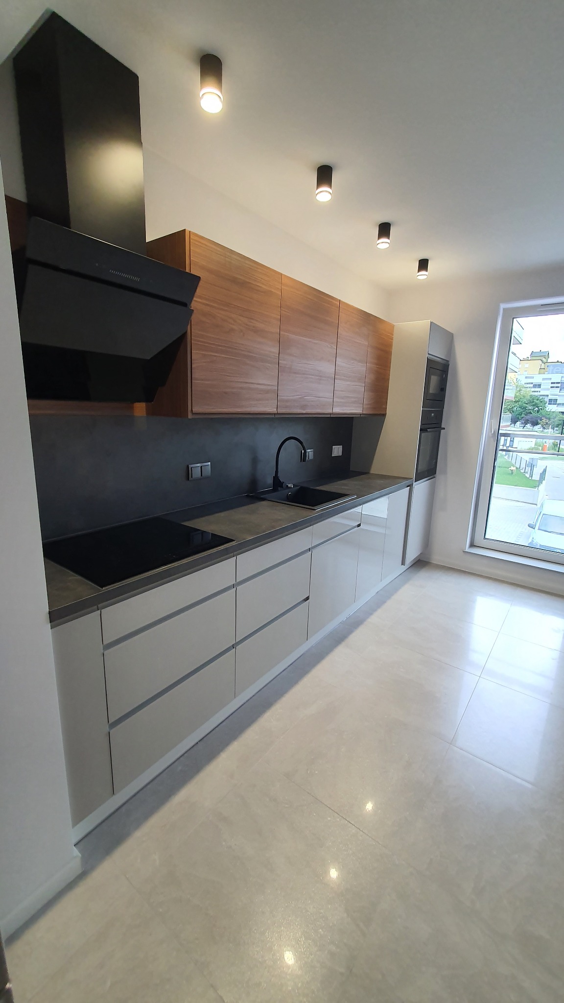 Nowoczesna kuchnia z drewnianymi szafkami górnymi i jasnymi dolnymi, czarnym okapem i płytą indukcyjną. Widok na okno z zielenią na zewnątrz.