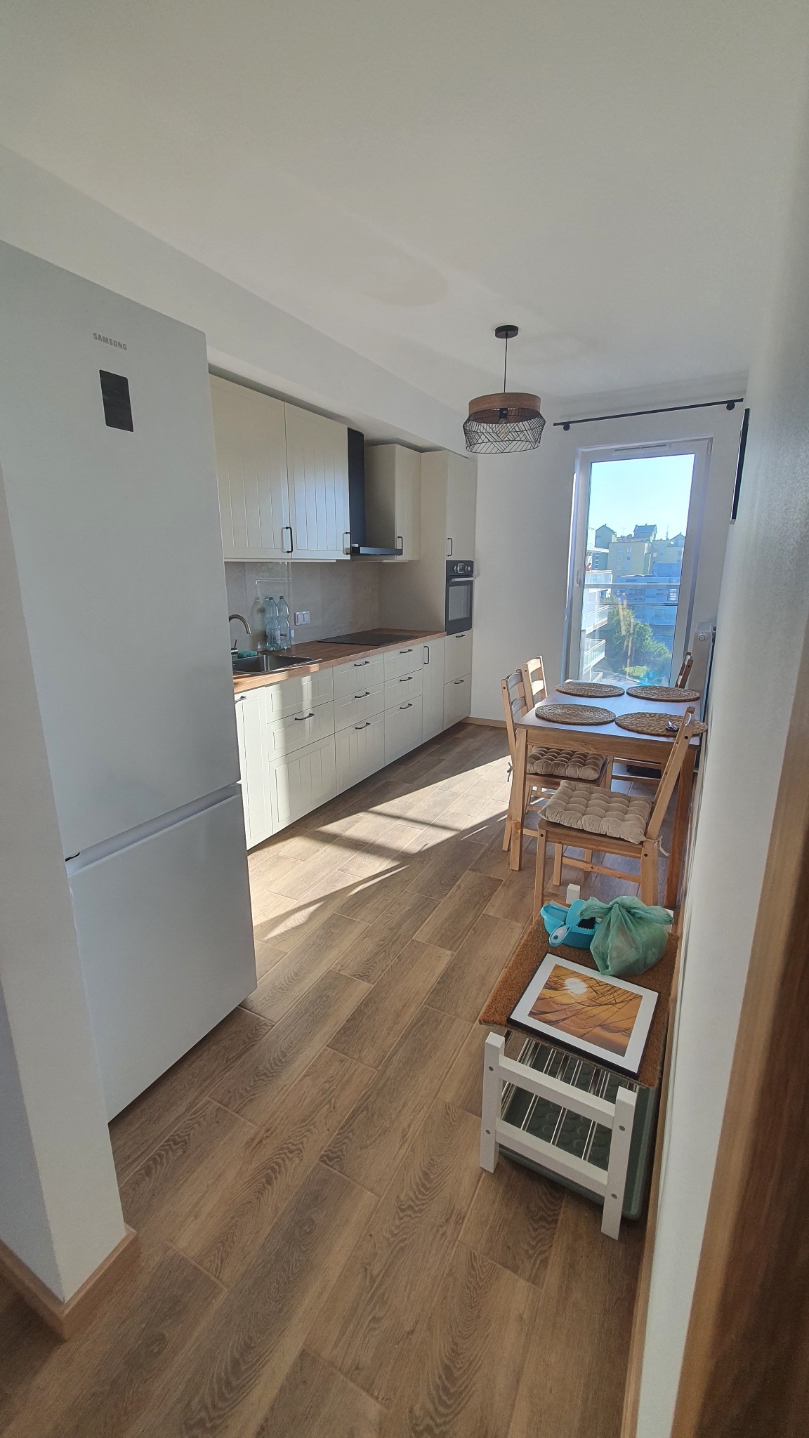 Jasna kuchnia z drewnianą podłogą i stołem jadalnym przy oknie. Widoczne białe szafki kuchenne, lodówka Samsung i dekoracyjny żyrandol.