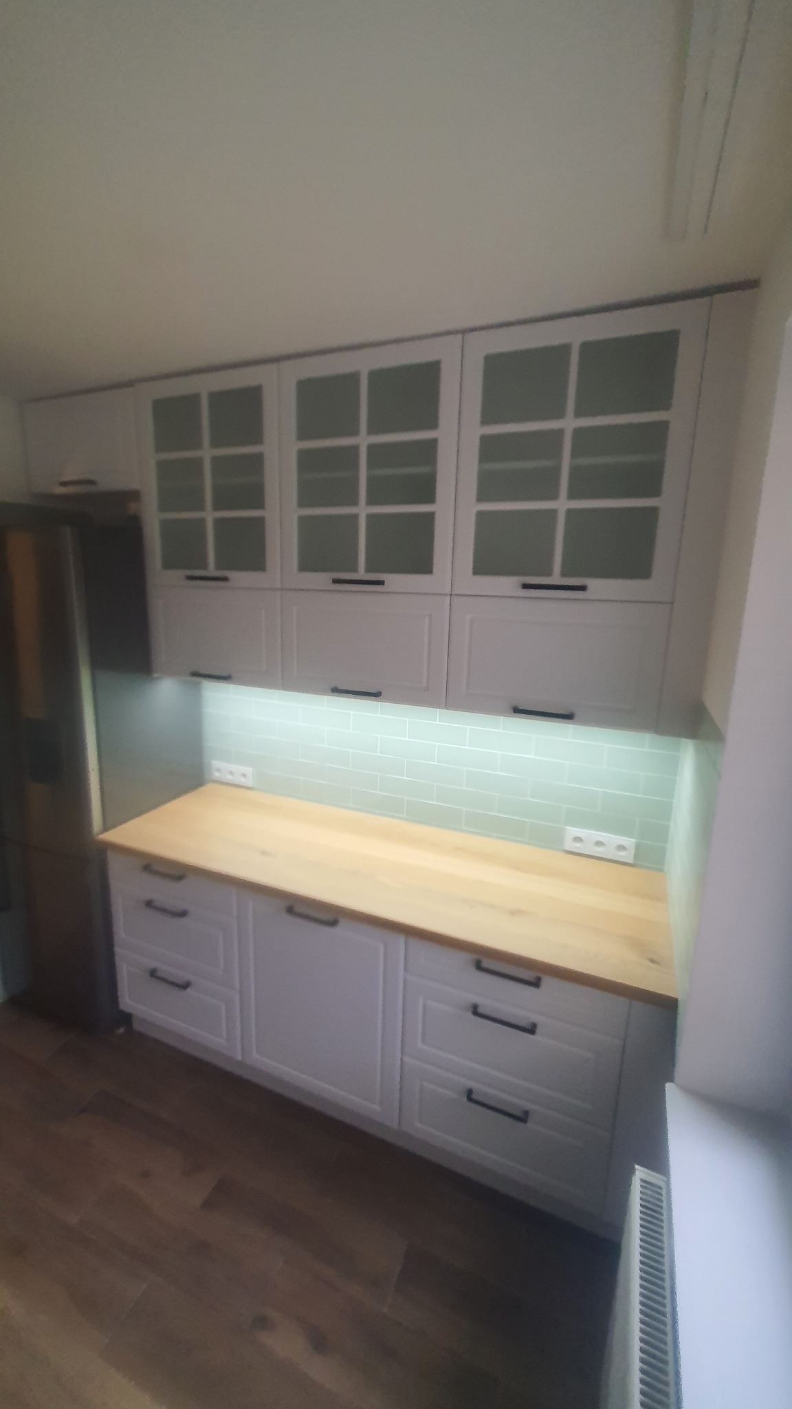 Jasna kuchnia z drewnianym blatem i białymi szafkami z przeszklonymi frontami. Podświetlenie LED nad blatem. Nowoczesny design z ciemnymi uchwytami.