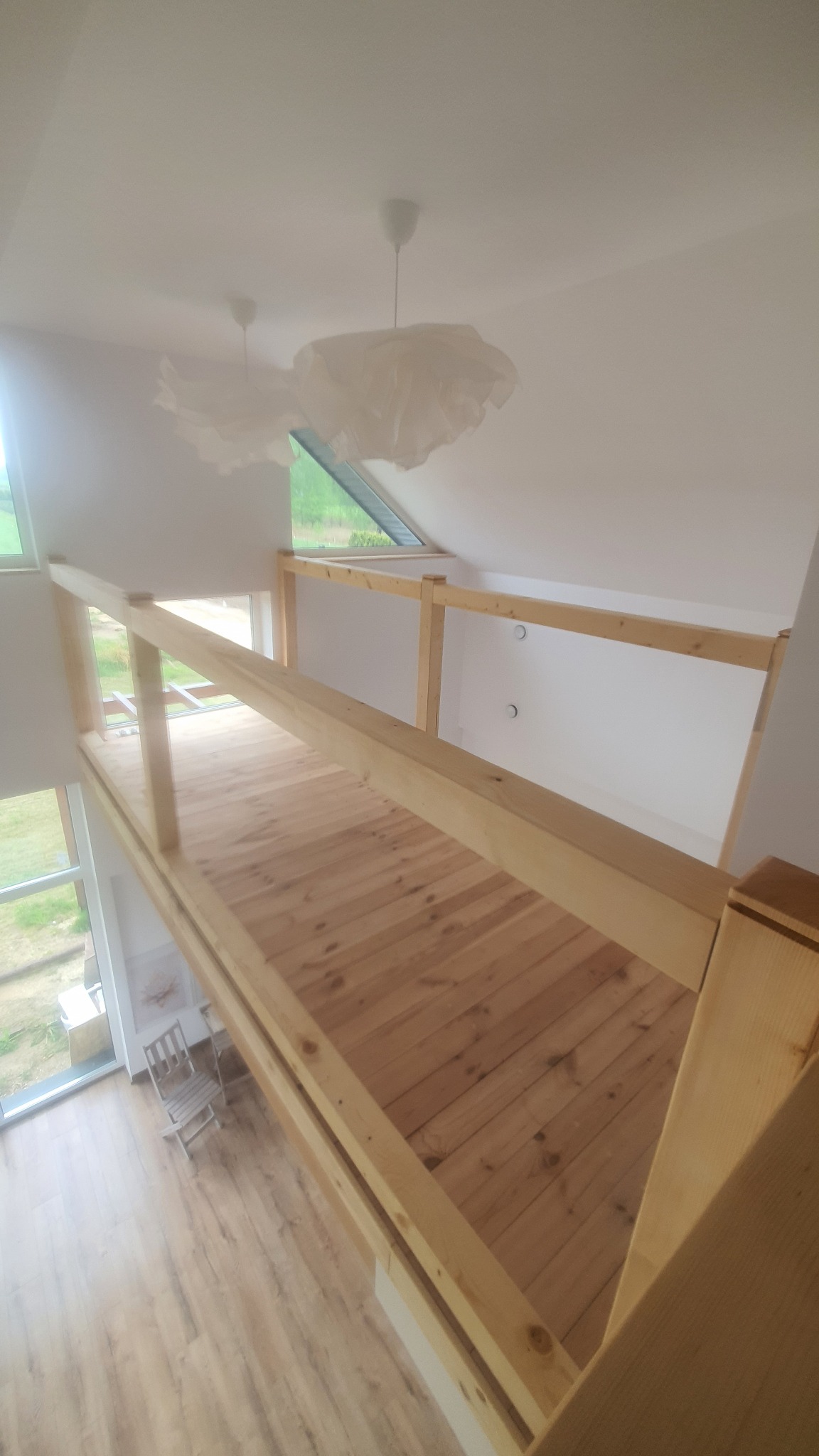 Drewniana antresola z balustradą w jasnym wnętrzu. Widok z góry na drewnianą podłogę i białe ściany. Dwa białe abażury w tle. Widoczne okno dachowe i podłoga na dole.