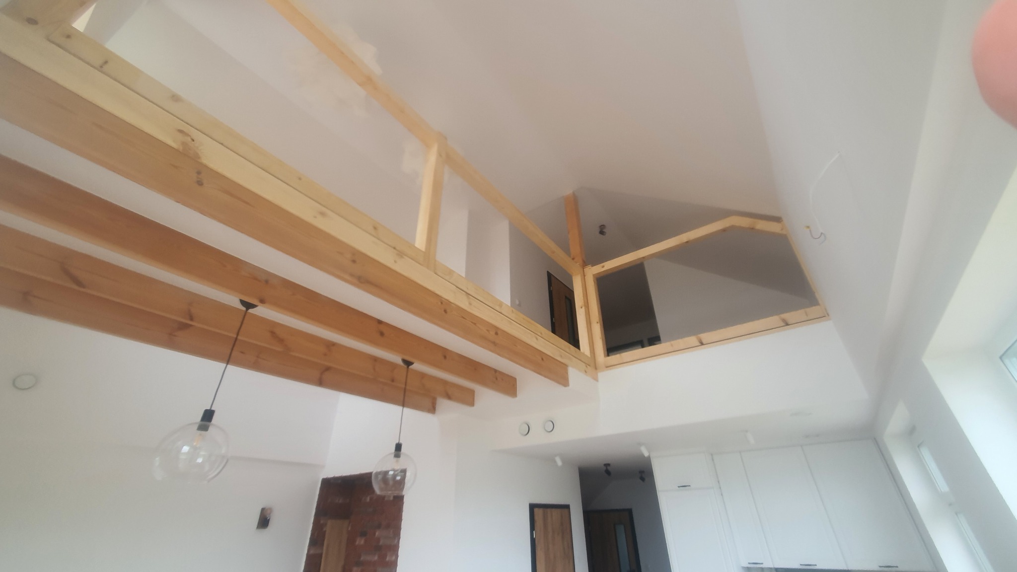 Drewniana konstrukcja antresoli w jasnym wnętrzu z białymi ścianami i widocznymi belkami stropowymi. Nowoczesne lampy wiszące i fragment zabudowy kuchennej w tle.