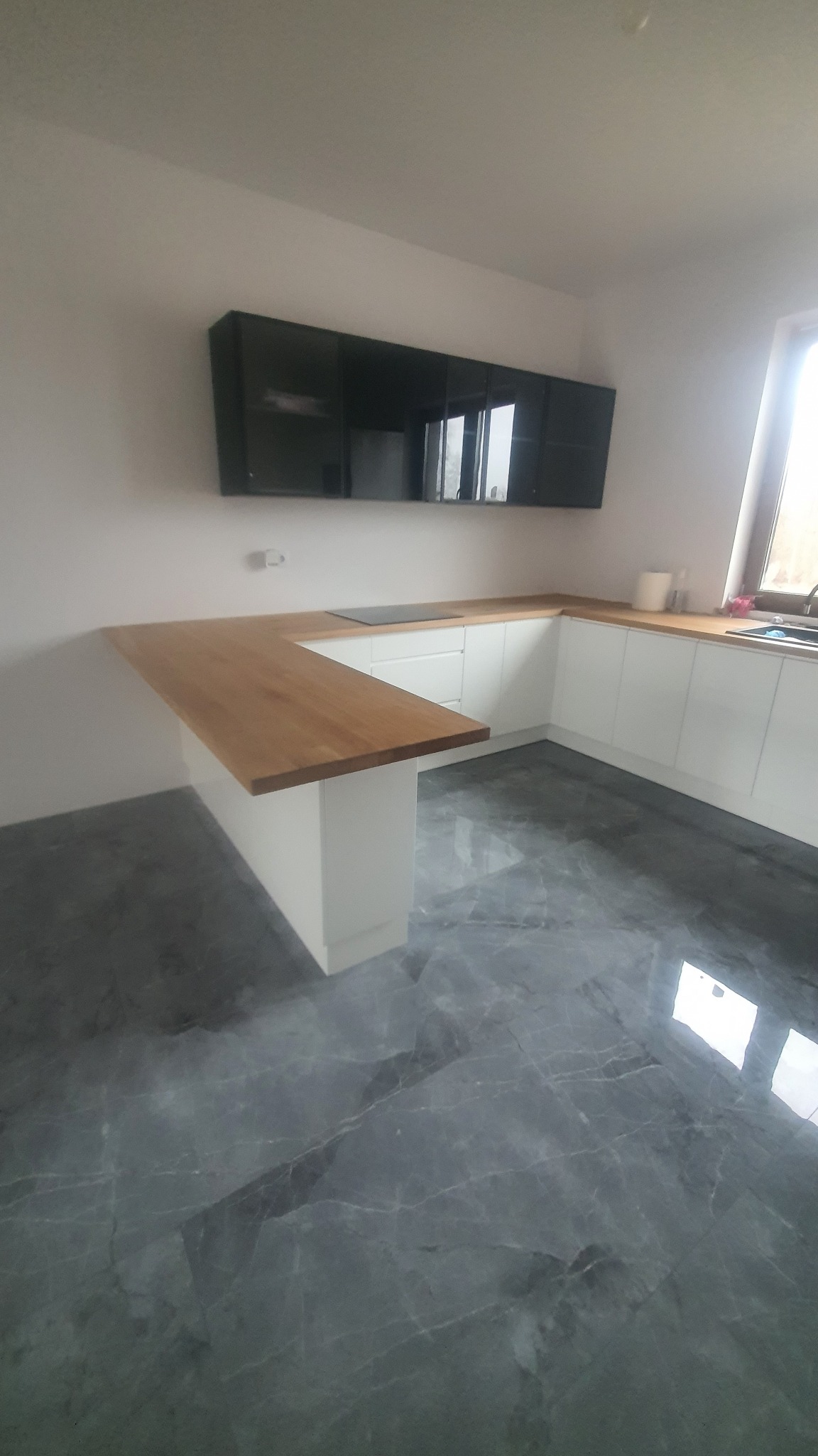 Nowoczesna kuchnia z białymi szafkami, drewnianym blatem i szarymi marmurowymi płytkami podłogowymi. Czarne szafki wiszące dodają elegancji.