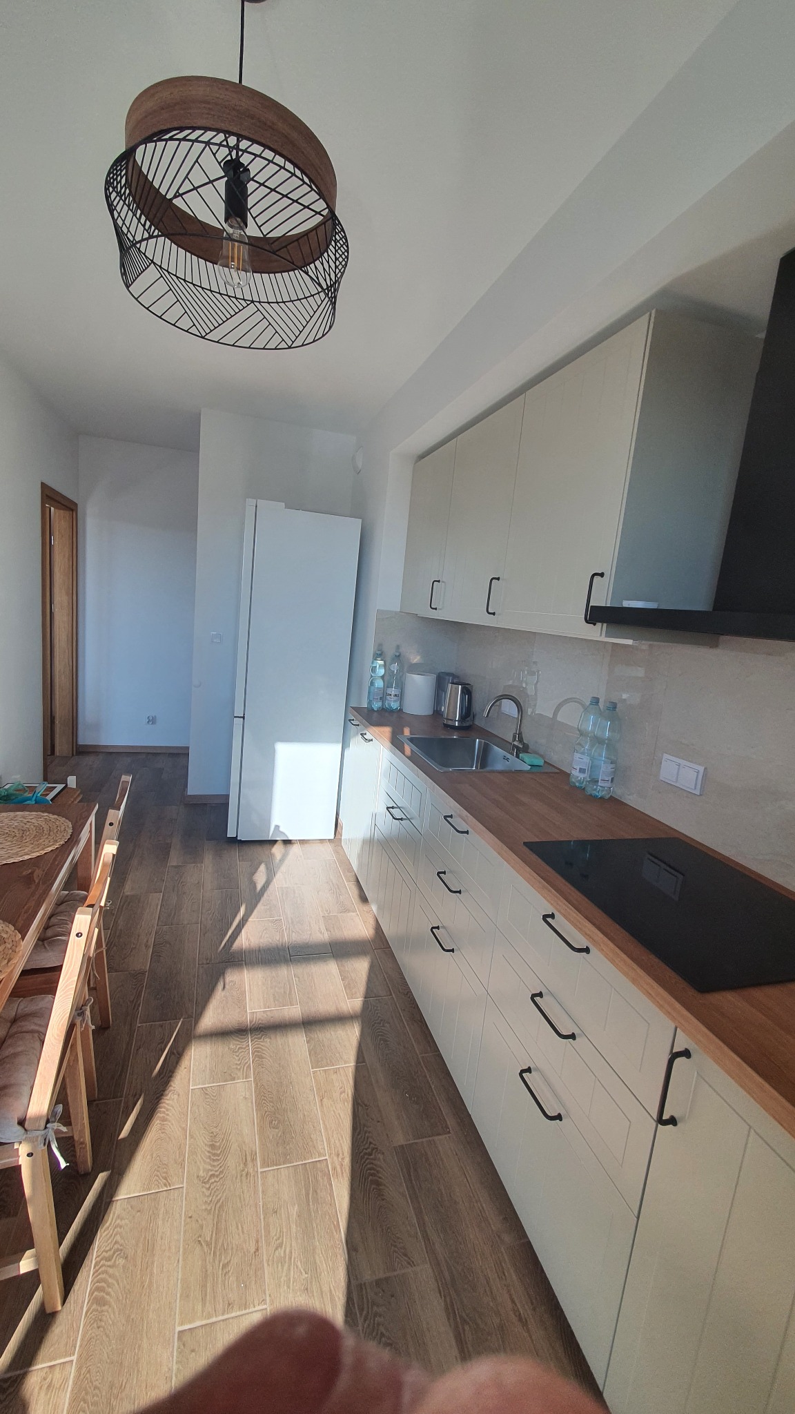 Jasna kuchnia z drewnianym blatem i podłogą imitującą drewno. Nowoczesna lampa sufitowa. Widok na lodówkę i drzwi do innego pomieszczenia. Ujęcie z dołu.