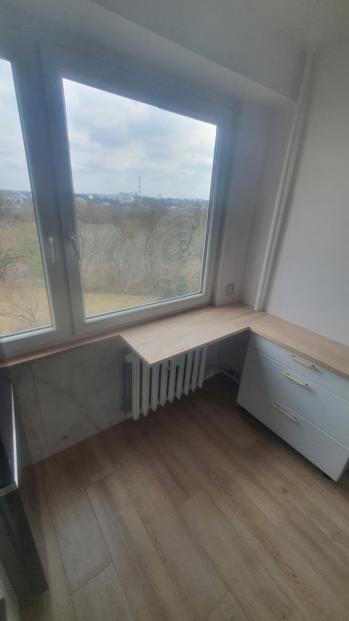 Białe okno z drewnianym blatem nad grzejnikiem i szafką z szufladami. Widok na zewnątrz z drzewami i budynkami w tle. Jasne wnętrze z drewnianą podłogą.