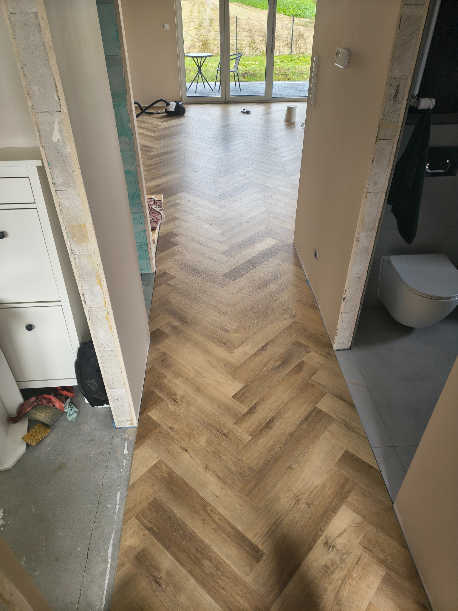 Ułożony parkiet w jodełkę przechodzi przez otwarte drzwi do łazienki z toaletą. Widoczne niedokończone ściany i podłoga przedpokoju. W tle okno z widokiem na zewnątrz.