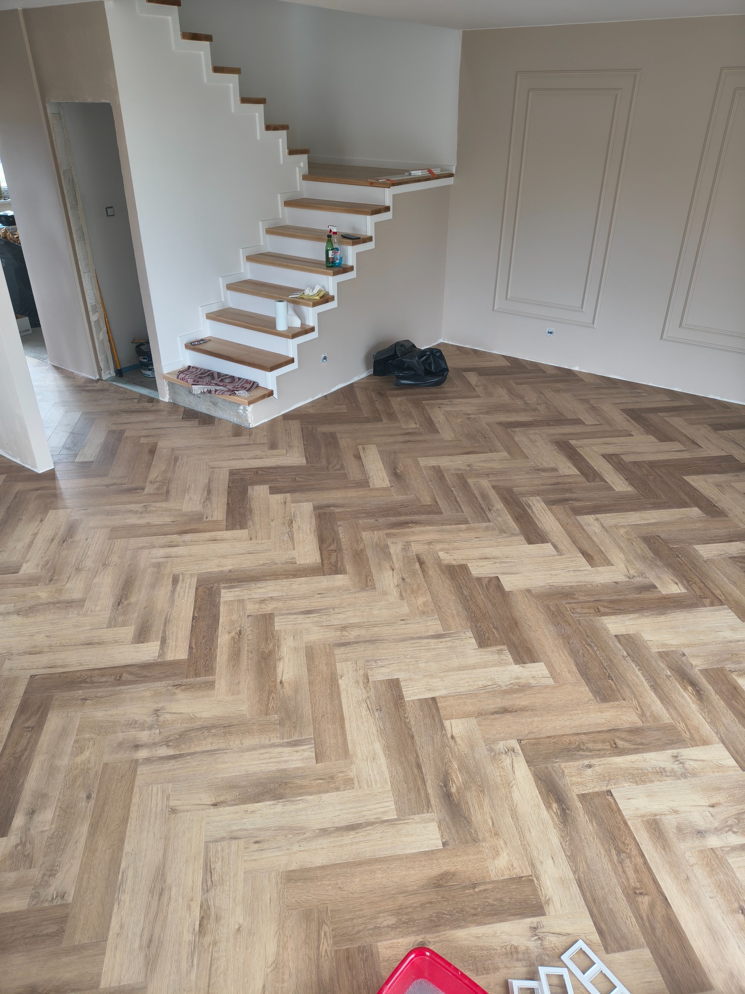 Ukończona podłoga z paneli ułożonych w jodełkę, jasne ściany z dekoracyjnymi panelami, schody z drewnianymi stopniami. Widoczne akcesoria montażowe.