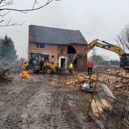 WJK INFRASTRUKTURA SPÓŁKA Z OGRANICZONĄ ODPOWIEDZIALNOŚCIĄ - Rozbiórka ceglanego domu z użyciem ciężkiego sprzętu CAT. Na pierwszym planie ognisko i błotniste podłoże. Dwóch pracowników nadzoruje prace. Pochmurny dzień.