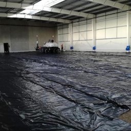 WJK INFRASTRUKTURA SPÓŁKA Z OGRANICZONĄ ODPOWIEDZIALNOŚCIĄ - Wnętrze hali w trakcie budowy: rozłożona czarna folia izolacyjna na podłożu, w tle maszyna do rozprowadzania izolacji i pracownik. Widoczne stalowe elementy konstrukcyjne.