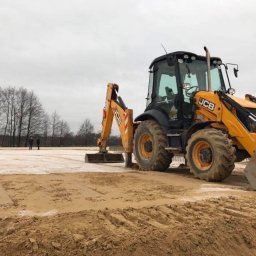 WJK INFRASTRUKTURA SPÓŁKA Z OGRANICZONĄ ODPOWIEDZIALNOŚCIĄ - Żółta koparko-ładowarka JCB na placu budowy z przygotowanym podłożem. W tle drzewa i dwie osoby. Prace ziemne w toku, przygotowanie terenu pod budowę.