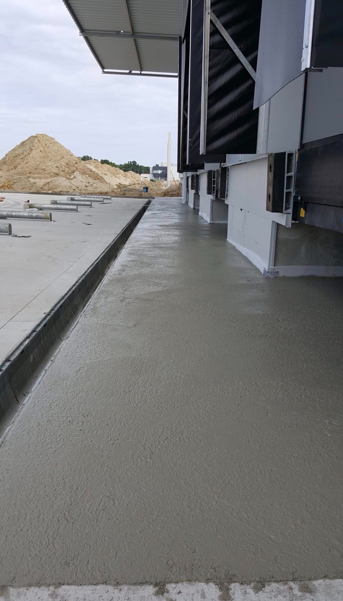 Wylana posadzka przemysłowa z odpływem liniowym w Małuszynie. Widoczny fragment hali i hałda piasku w tle. Świeży beton, równa powierzchnia.
