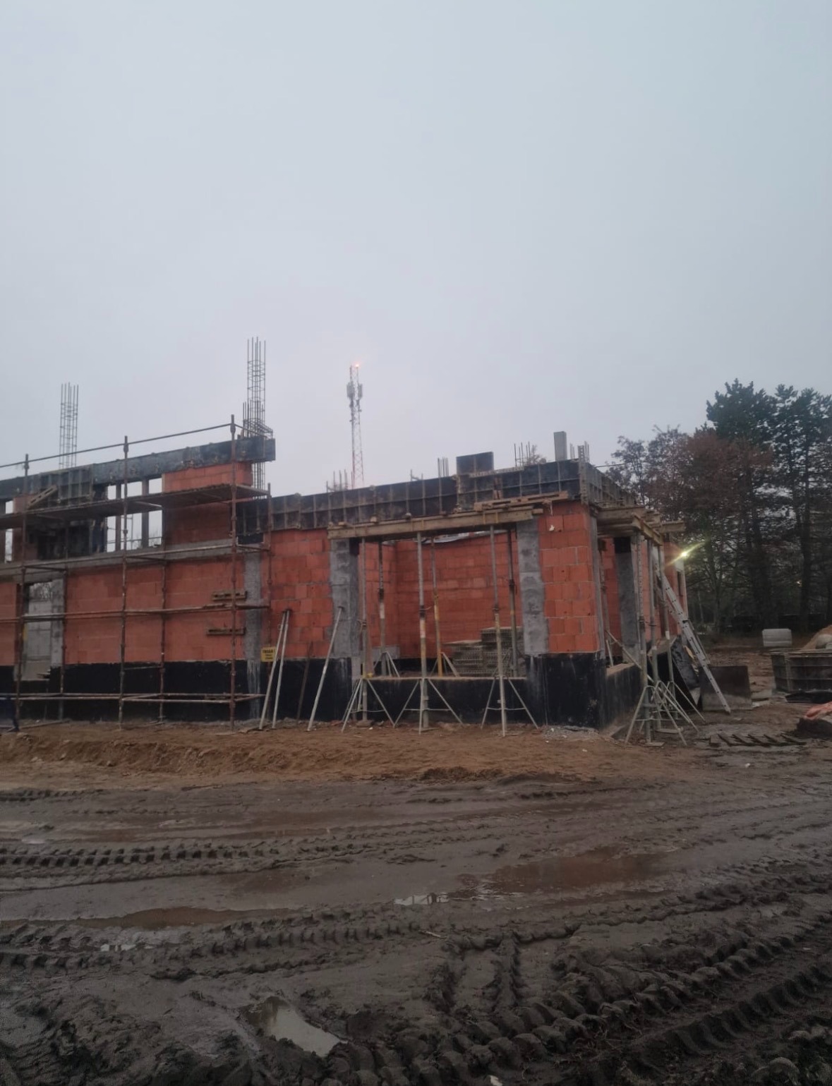 Wznoszenie ścian z czerwonej cegły na fundamencie, z widocznym szalunkiem i rusztowaniem. W tle drzewa i maszt telekomunikacyjny. Błotniste podłoże.