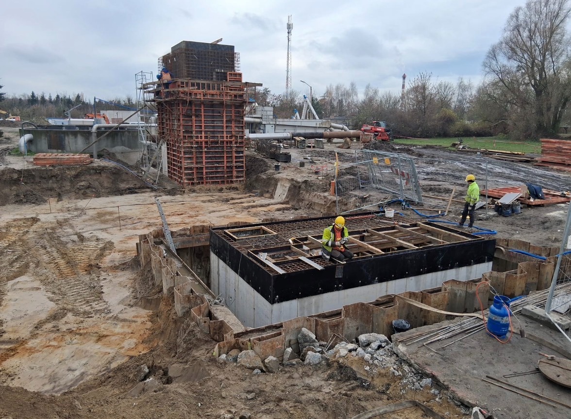 Budowa zbiornika żelbetowego: pracownik w żółtym kasku siedzi na zbrojeniu, w tle widoczna konstrukcja szalunkowa i teren budowy z maszynami. Pochmurny dzień.