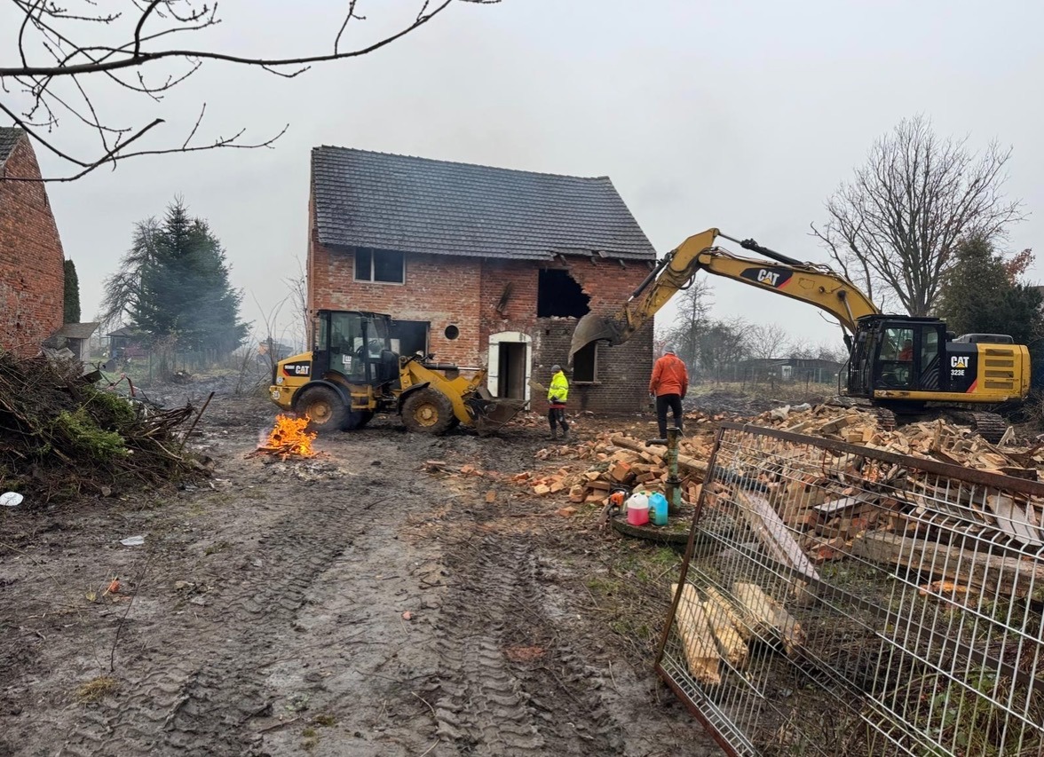Rozbiórka ceglanego domu z użyciem ciężkiego sprzętu CAT. Na pierwszym planie ognisko i błotniste podłoże. Dwóch pracowników nadzoruje prace. Pochmurny dzień.