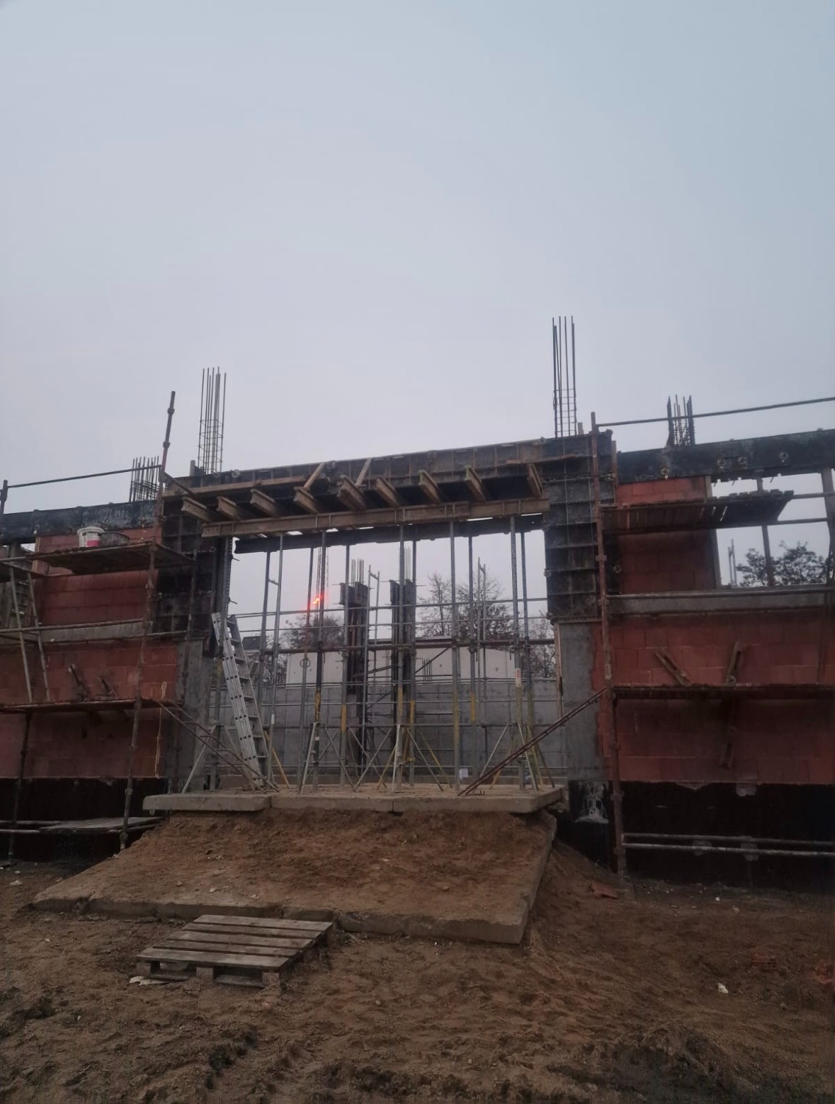 Wznoszenie konstrukcji żelbetowej: szalunki, zbrojenie i cegła klinkierowa tworzą surowy, industrialny krajobraz budowy. Widoczne rusztowania i drabina.