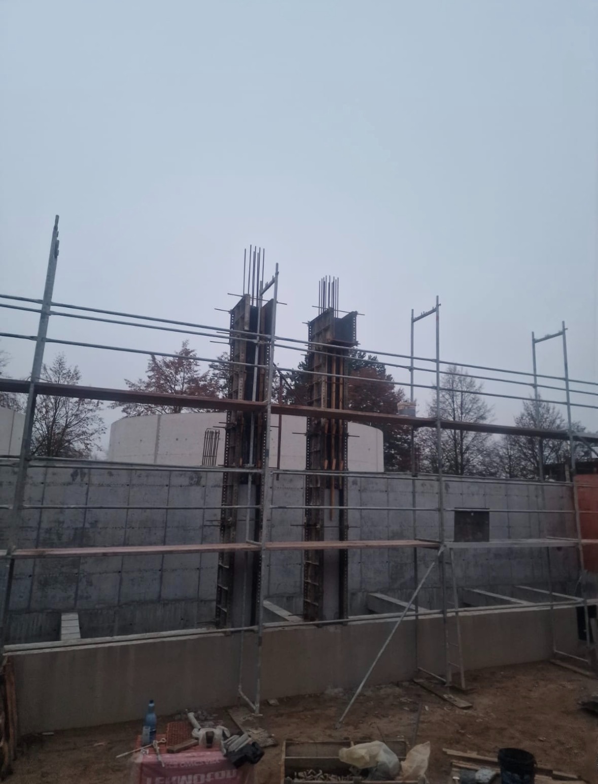 Żelbetowa konstrukcja w trakcie budowy, widoczne szalunki kolumn i ściany fundamentowe, rusztowania. Pochmurne niebo w tle.