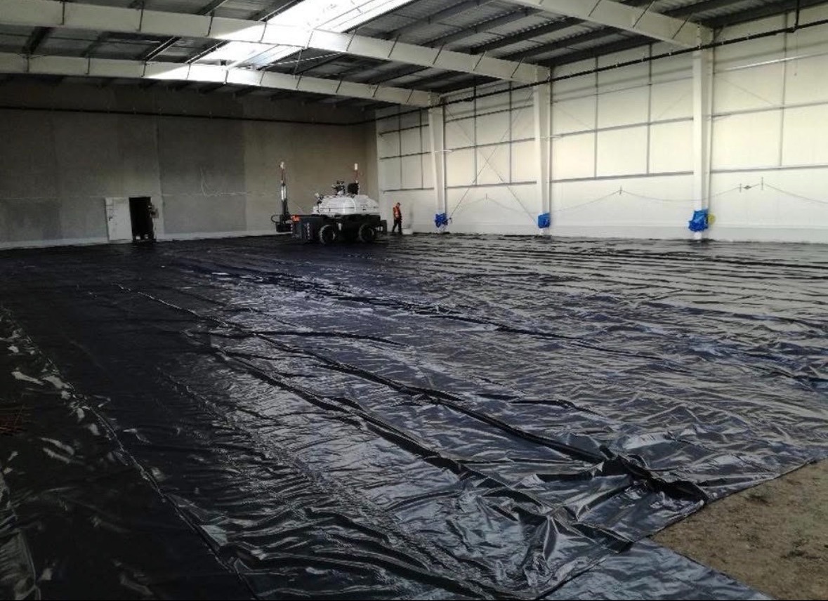 Wnętrze hali w trakcie budowy: rozłożona czarna folia izolacyjna na podłożu, w tle maszyna do rozprowadzania izolacji i pracownik. Widoczne stalowe elementy konstrukcyjne.