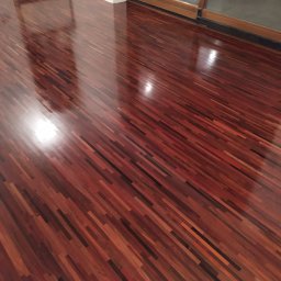 PPHU EUROPARKIET - Błyszcząca, świeżo wycyklinowana podłoga z egzotycznego drewna w odcieniach czerwieni i brązu, ułożona w parkiet. Widoczne refleksy światła i fragment okna balkonowego.