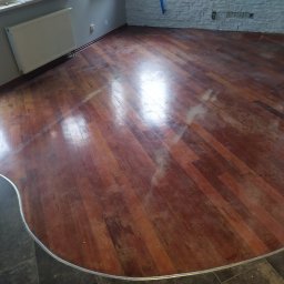 PPHU EUROPARKIET - Błyszcząca, odnowiona podłoga z parkietu ułożonego w jodełkę, z widocznymi śladami po cyklinowaniu, przechodząca w ciemną, kamienną posadzkę przy ścianie.