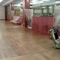 PPHU EUROPARKIET - Cyklinowanie parkietu w przestronnym biurze. Parkiet z wyraźnym wzorem, zabezpieczone meble, pracownik z maszyną do cyklinowania. Widoczne ślady po cyklinowaniu.