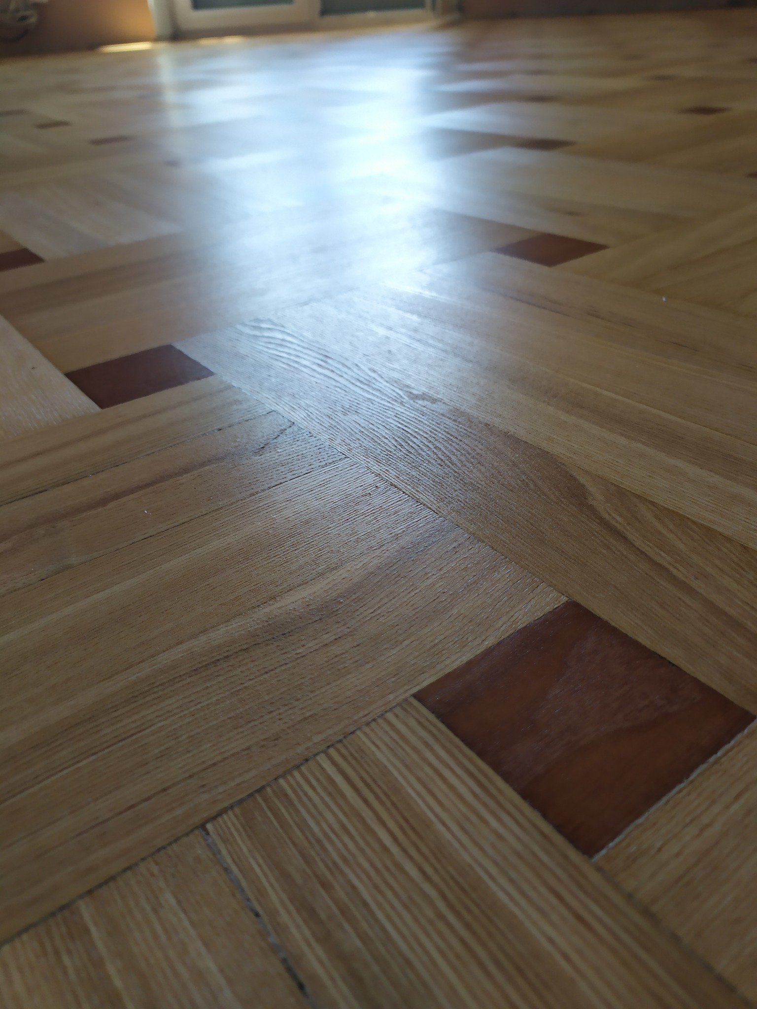 Ułożony parkiet w jodełkę z ciemniejszymi wstawkami, wykończony lakierem z połyskiem. Widoczne naturalne usłojenie drewna i odbicie światła od podłogi.