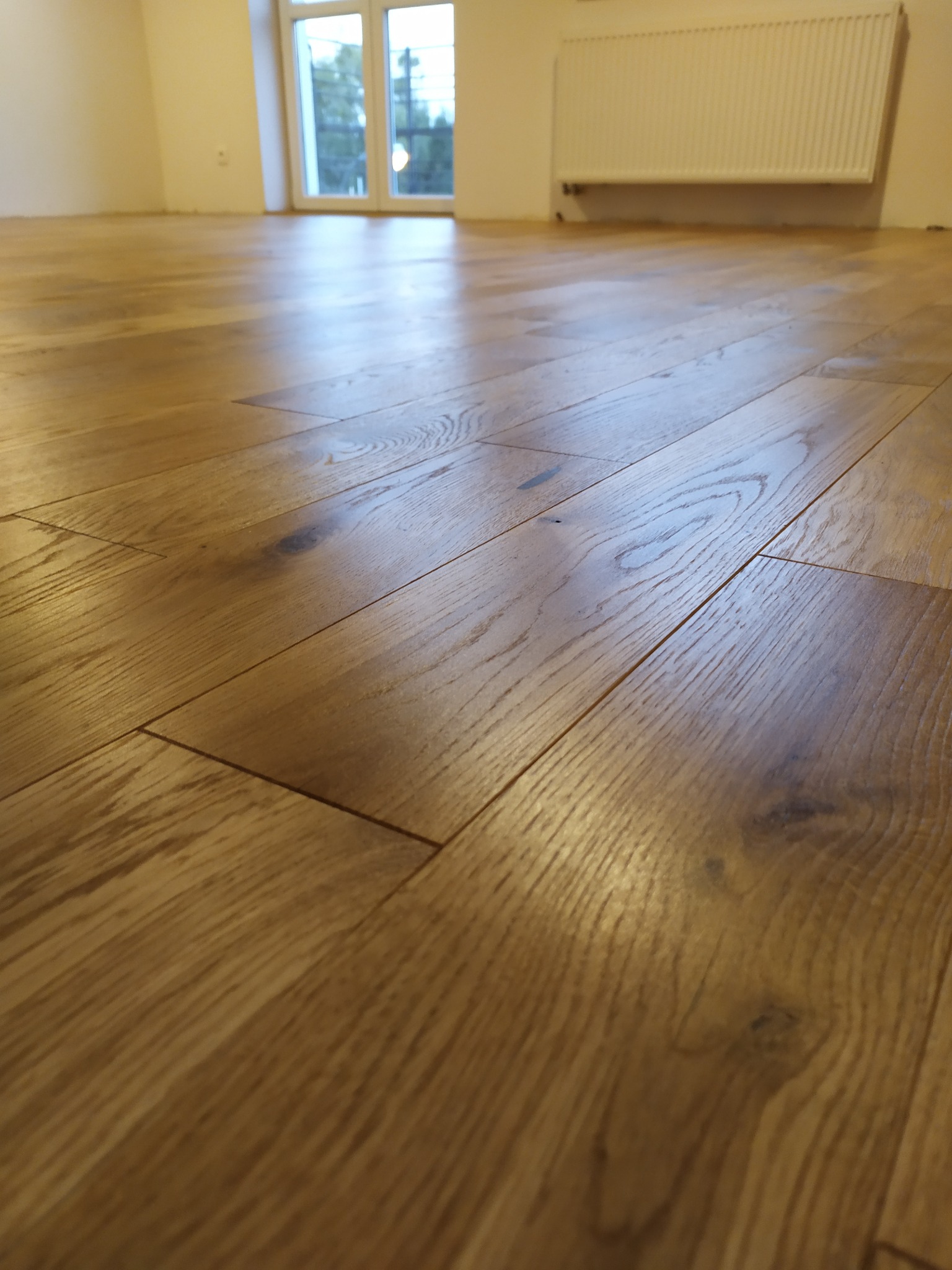 Jasny, drewniany parkiet ułożony w nowym pomieszczeniu z oknem i grzejnikiem. Widok z niskiej perspektywy podkreśla strukturę drewna i precyzję ułożenia.