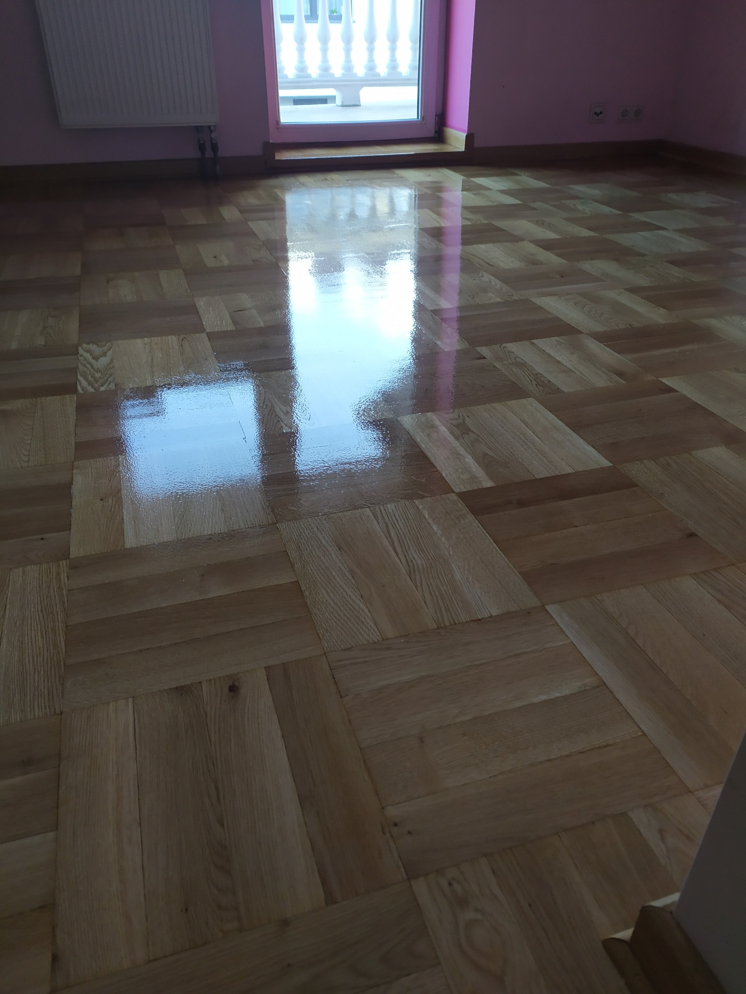 Błyszcząca, odnowiona podłoga parkietowa ułożona w kwadraty, odbijająca światło okna balkonowego w pokoju z różowymi ścianami i grzejnikiem.