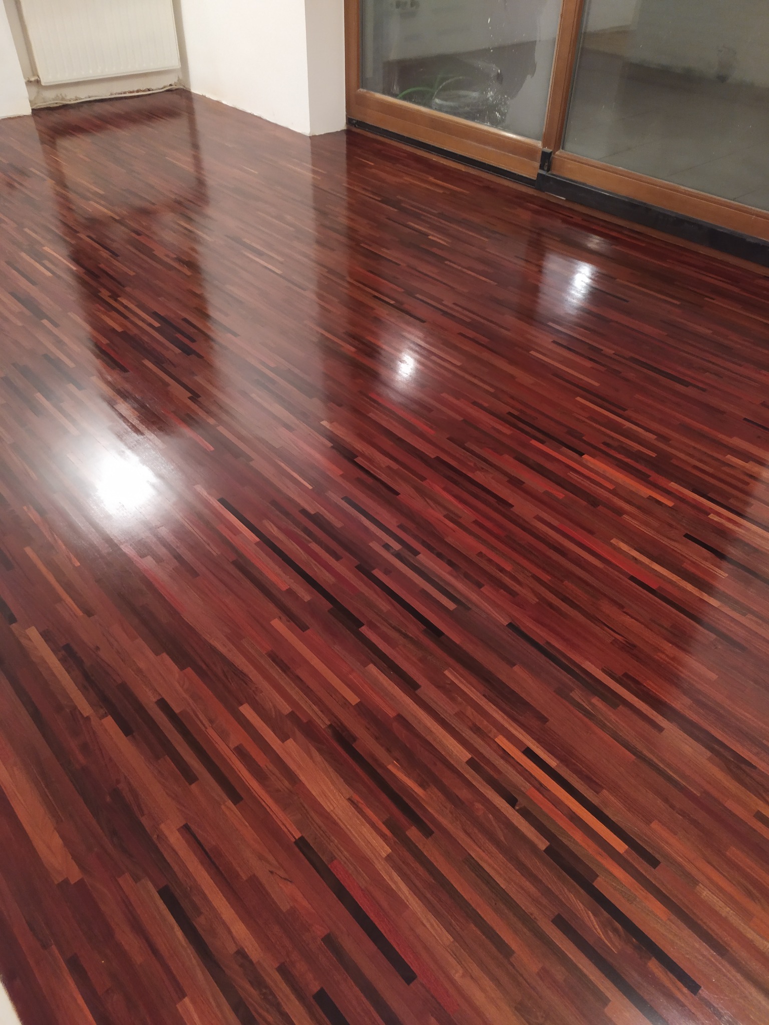 Błyszcząca, świeżo wycyklinowana podłoga z egzotycznego drewna w odcieniach czerwieni i brązu, ułożona w parkiet. Widoczne refleksy światła i fragment okna balkonowego.