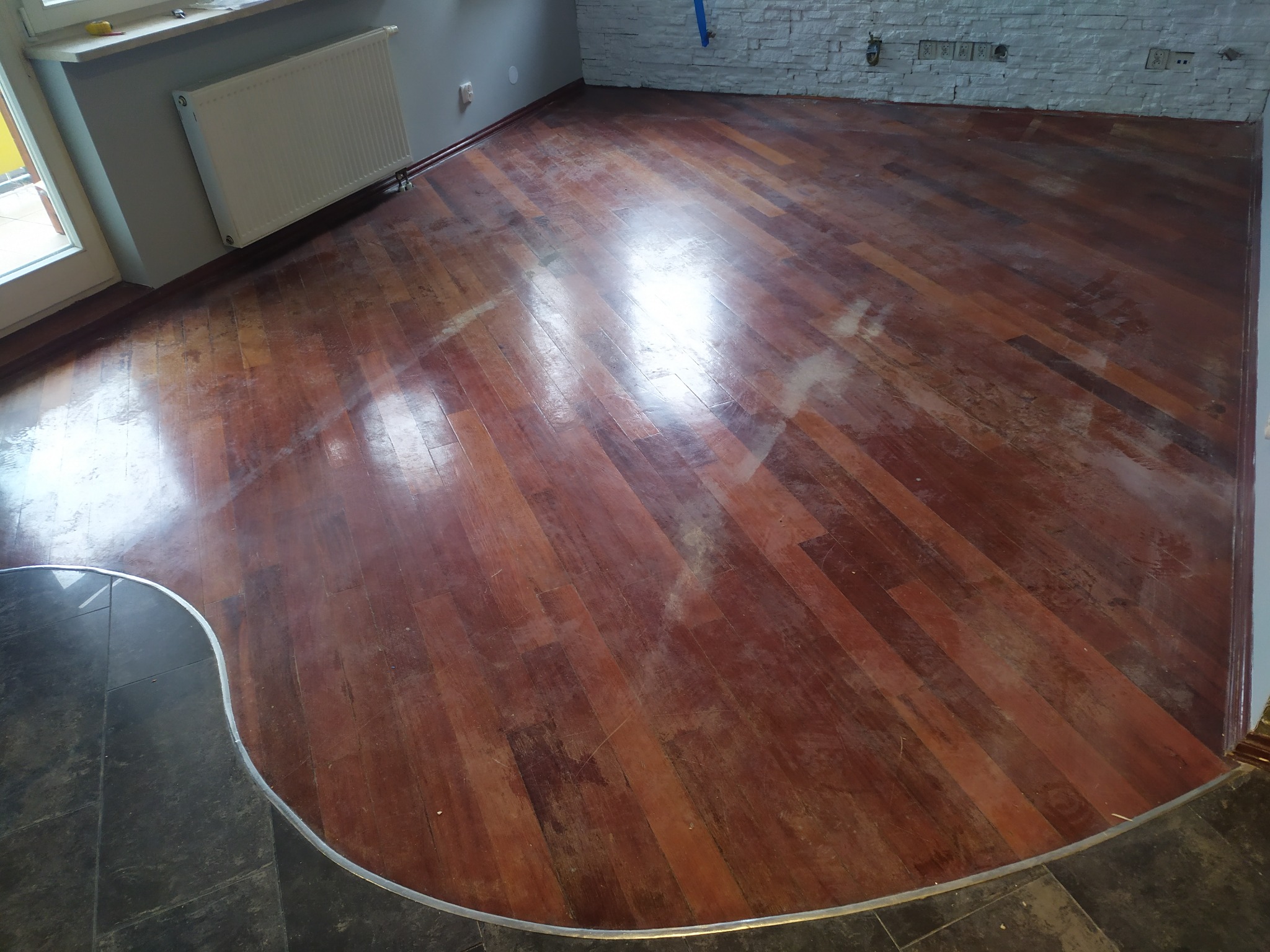 Błyszcząca, odnowiona podłoga z parkietu ułożonego w jodełkę, z widocznymi śladami po cyklinowaniu, przechodząca w ciemną, kamienną posadzkę przy ścianie.