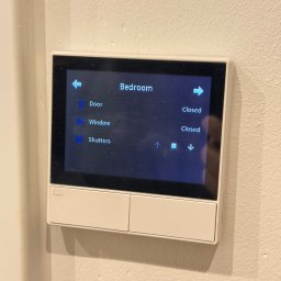 eHelper.eu Jordan Jasiński - Panel sterowania inteligentnego domu na ścianie, z ekranem dotykowym pokazującym status zamkniętych drzwi, okien i rolet w sypialni. Widoczne strzałki do sterowania.