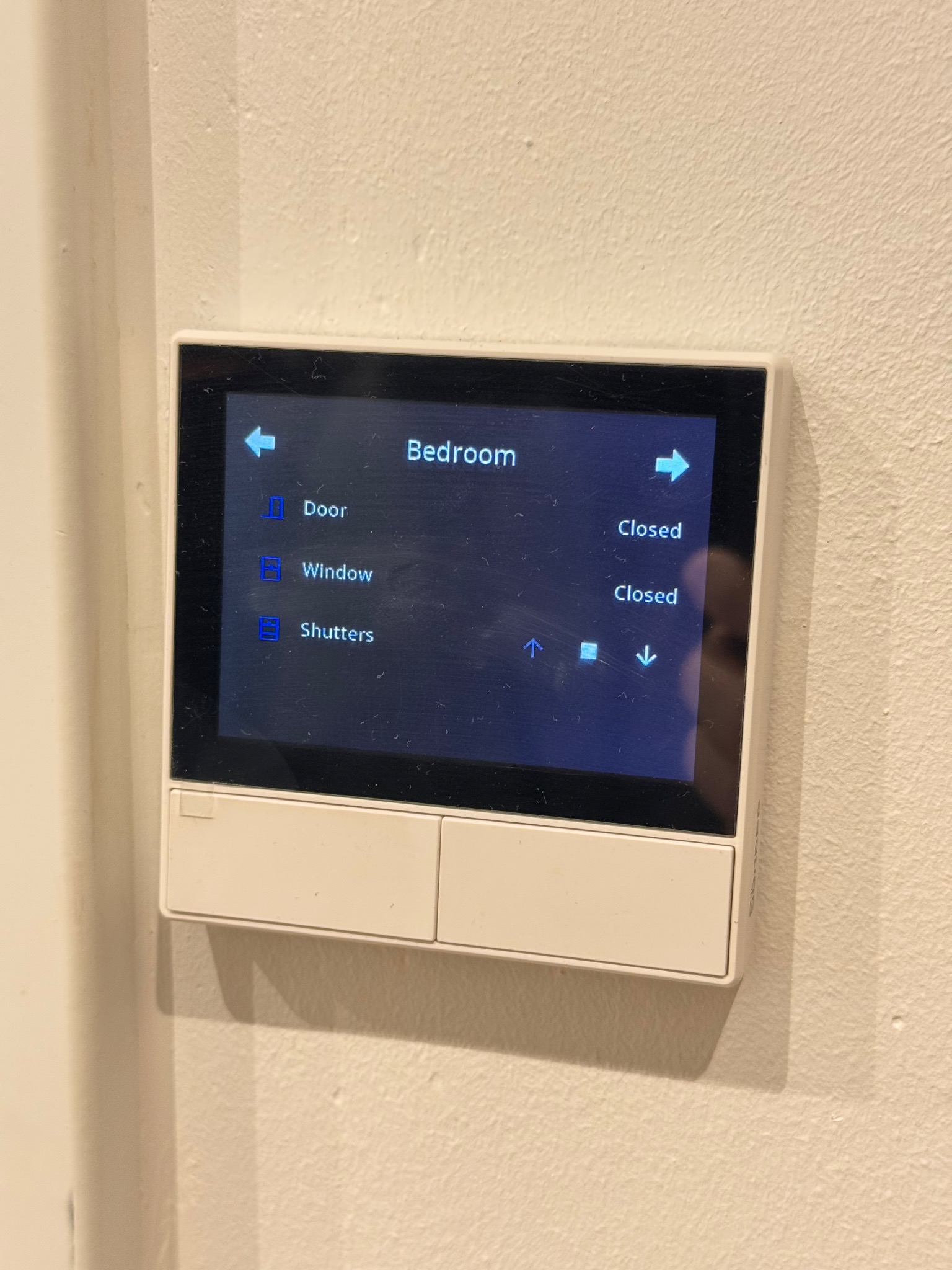 Panel sterowania inteligentnego domu na ścianie, z ekranem dotykowym pokazującym status zamkniętych drzwi, okien i rolet w sypialni. Widoczne strzałki do sterowania.