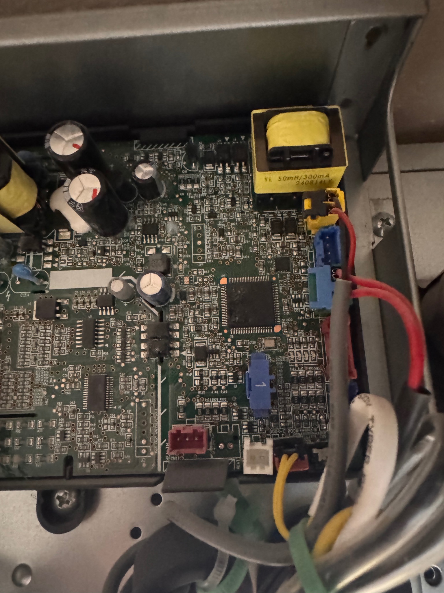 Płyta główna urządzenia elektronicznego w metalowej obudowie. Widoczne kondensatory, układy scalone, kolorowe wtyczki i przewody. Żółty transformator na górze.