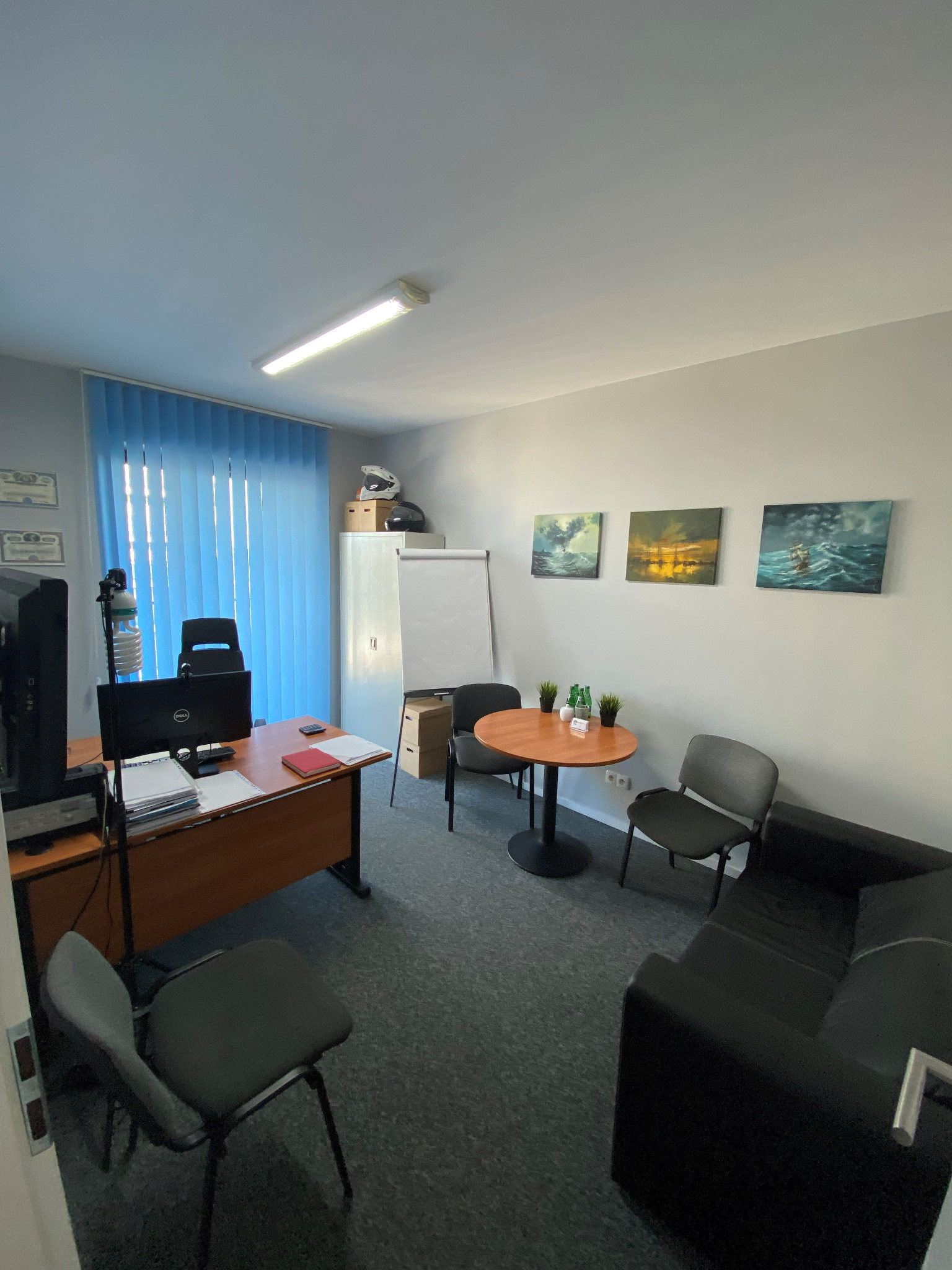 Jasne biuro z biurkiem, komputerem, krzesłami i sofą. Na ścianie wiszą obrazy z motywem morskim. W tle widoczne rolety i tablica suchościeralna.