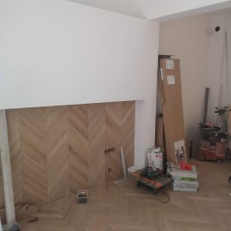 Remonty Wykończenia Tomasz Kulik - Ukończona podłoga z parkietu ułożonego w jodełkę francuską, z widocznymi narzędziami i materiałami budowlanymi w tle. Częściowo wykończona ściana.