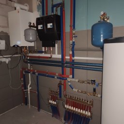 Remonty Wykończenia Tomasz Kulik - Złożona instalacja grzewcza z kotłem Kospel, rozdzielaczami i rurami w kolorach niebieskim i czerwonym, zamontowana w pomieszczeniu z szarą glazurą.