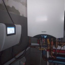 Remonty Wykończenia Tomasz Kulik - Nowoczesny, biały piec gazowy marki Termet z wyświetlaczem temperatury 38°C, podłączony do instalacji z rur miedzianych i kolorowych, na tle surowej ściany.