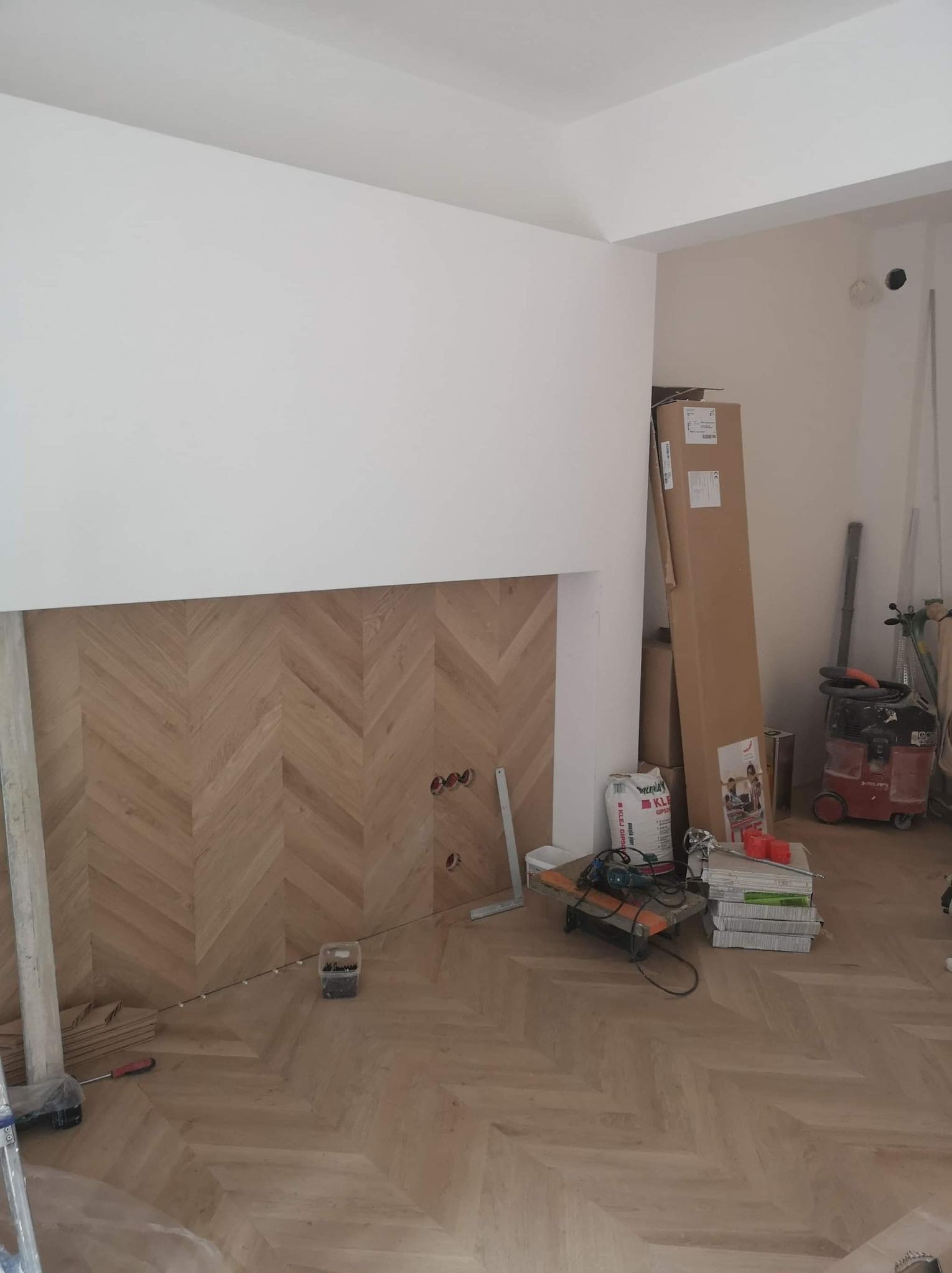 Ukończona podłoga z parkietu ułożonego w jodełkę francuską, z widocznymi narzędziami i materiałami budowlanymi w tle. Częściowo wykończona ściana.