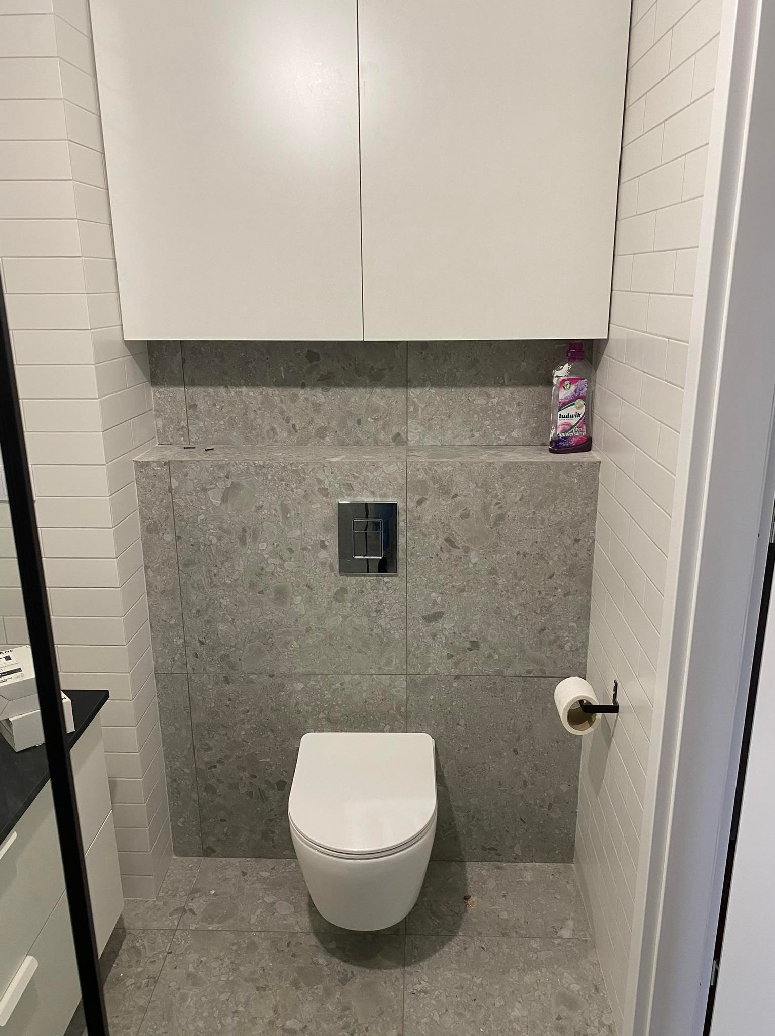 Nowoczesna toaleta z podwieszaną muszlą, szarą mozaiką i białymi płytkami. Nad toaletą szafka i butelka płynu. Minimalistyczny design, czyste linie, oświetlenie górne.