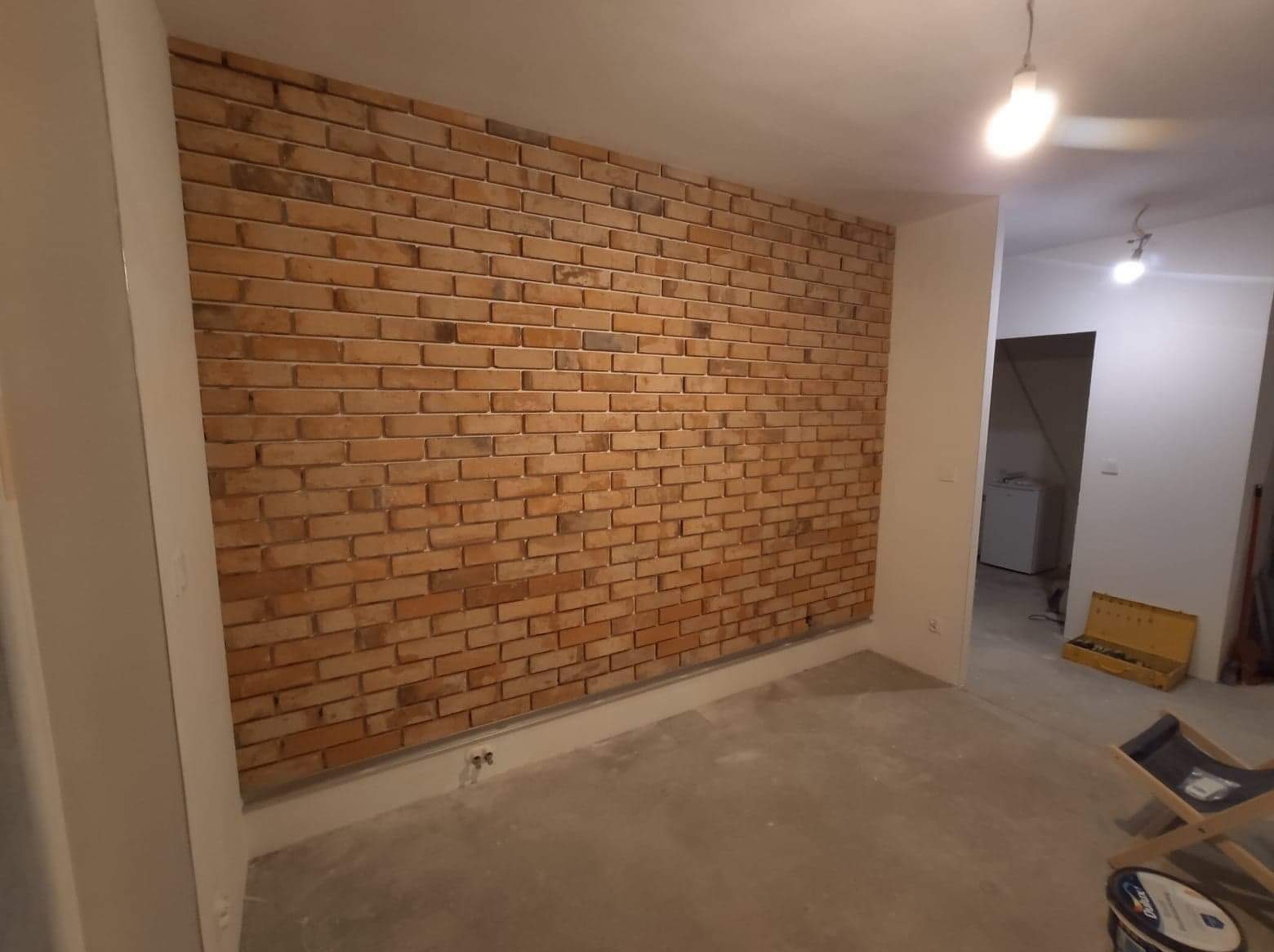 Ściana wykończona cegłą dekoracyjną w nowym wnętrzu. Widoczne narzędzia i farba, sugerujące etap wykończeniowy. Oświetlenie sufitowe podkreśla fakturę cegły.