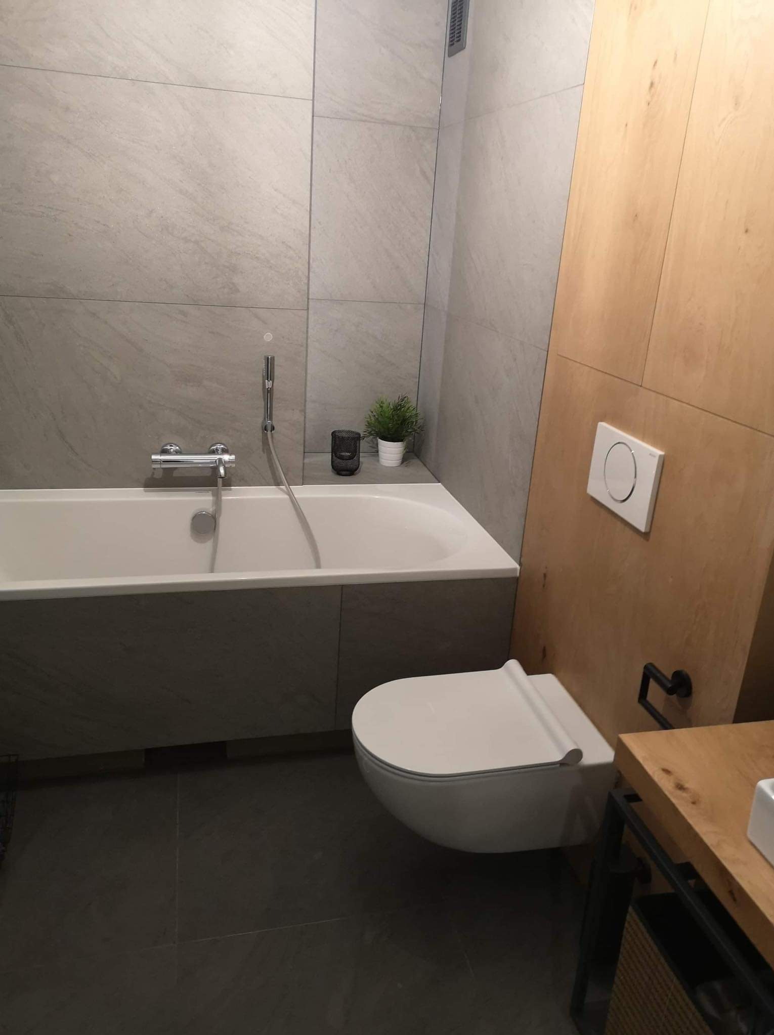 Nowoczesna łazienka z wanną, toaletą podwieszaną i szarą glazurą. Minimalistyczny design z drewnianymi akcentami i rośliną doniczkową.