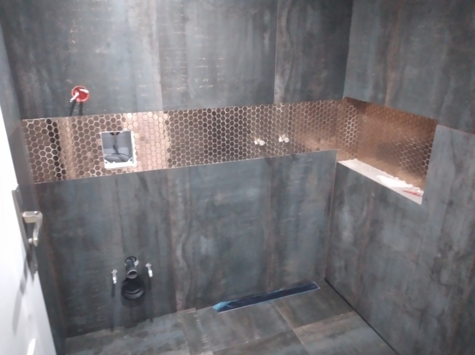 Łazienka w trakcie wykańczania w Pruszkowie: ciemne płytki imitujące metal, mozaika w odcieniach miedzi, widoczny odpływ liniowy i przygotowane punkty instalacyjne.