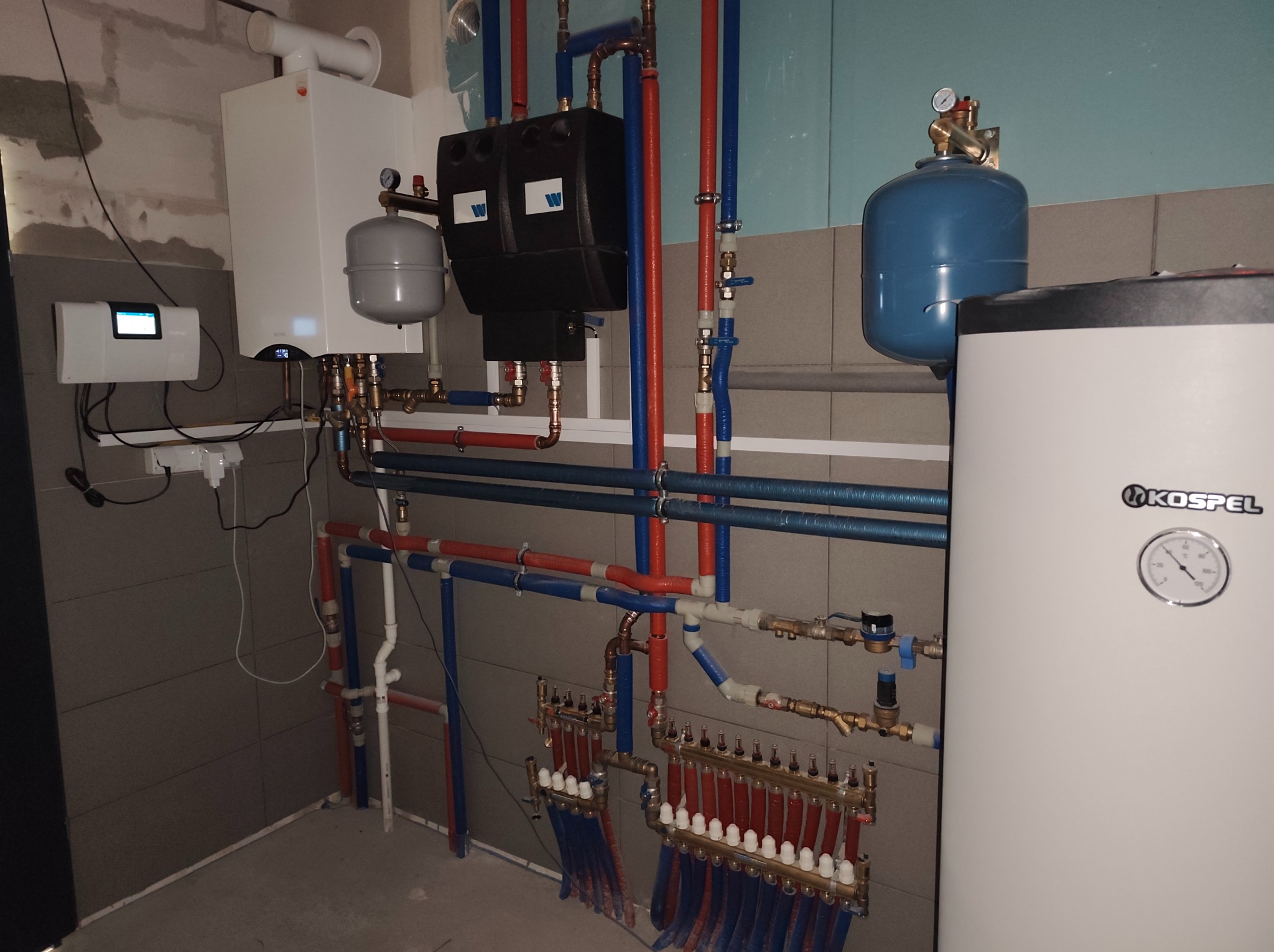 Złożona instalacja grzewcza z kotłem Kospel, rozdzielaczami i rurami w kolorach niebieskim i czerwonym, zamontowana w pomieszczeniu z szarą glazurą.
