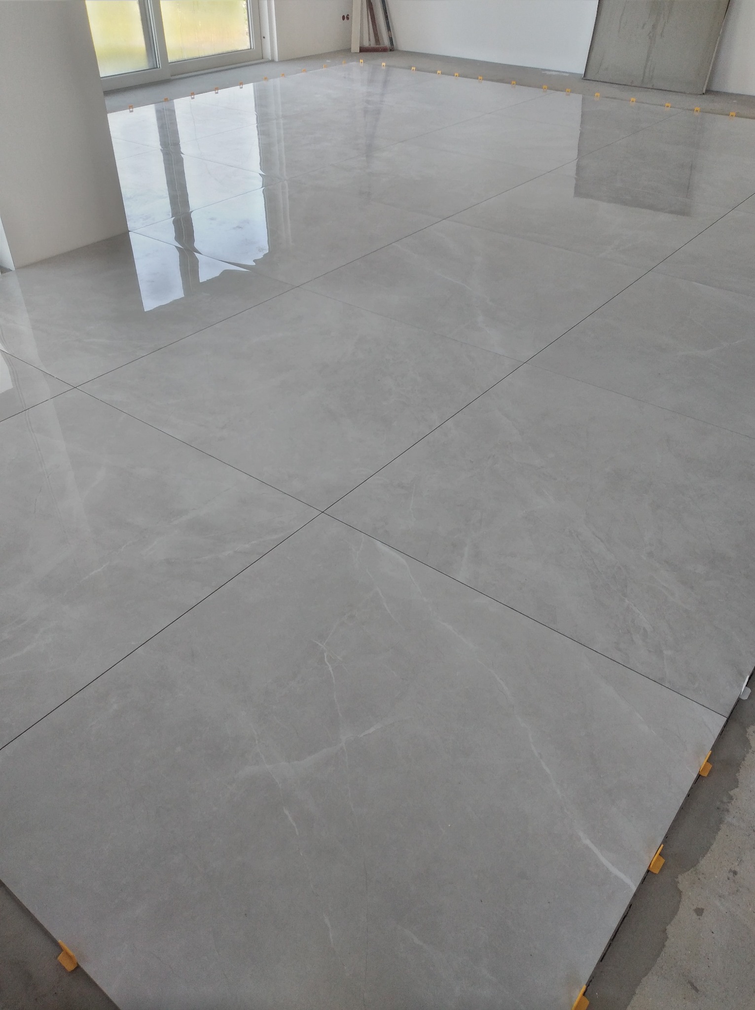 Połyskująca, szara podłoga z płytek gresowych w trakcie układania, z widocznymi żółtymi elementami poziomującymi. Duże formaty płytek, jasne wnętrze.