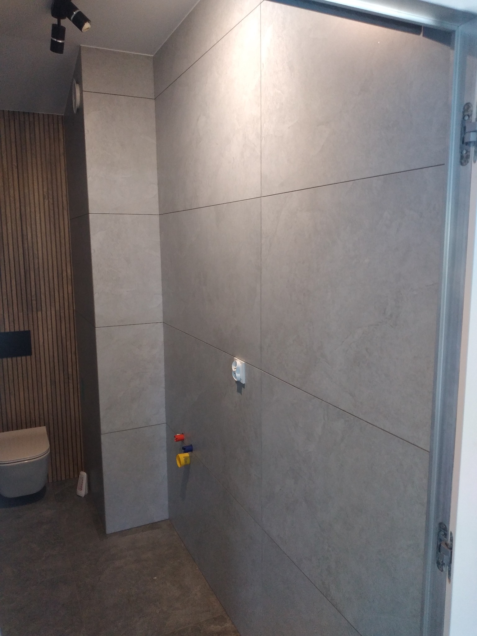 Wykończona łazienka z szarymi, matowymi płytkami wielkoformatowymi na ścianach i podłodze. Widoczne przyłącza wodne i częściowo toaleta. Minimalistyczne oświetlenie.