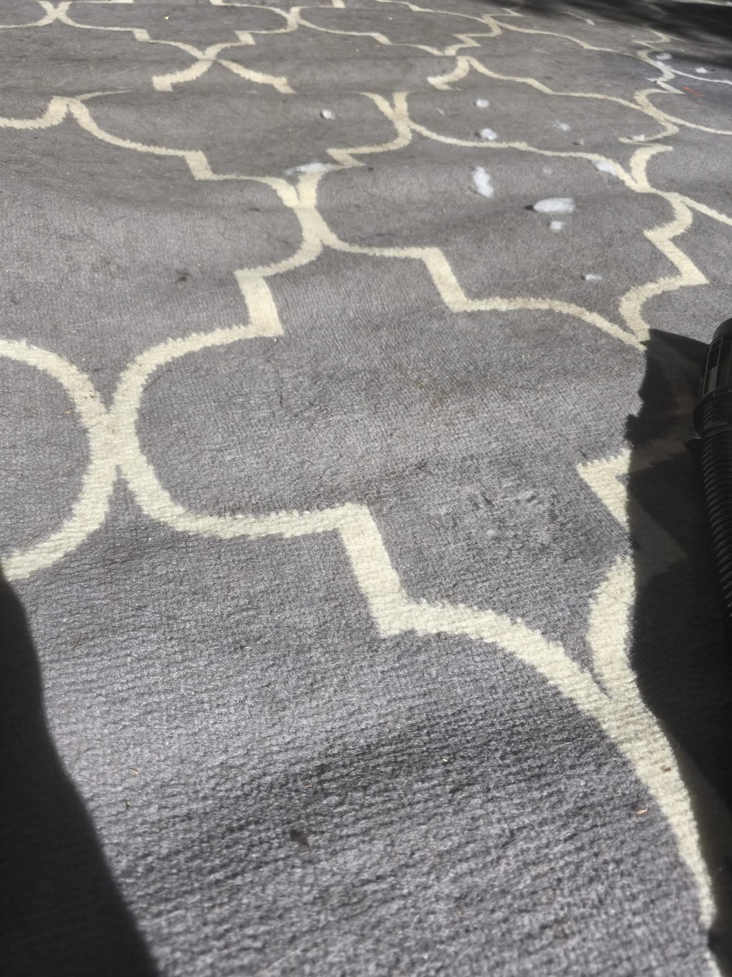 Szary dywan z geometrycznym wzorem w trakcie czyszczenia. Widoczne plamy z detergentu i fragment odkurzacza. Ujęcie z góry, ostre światło.