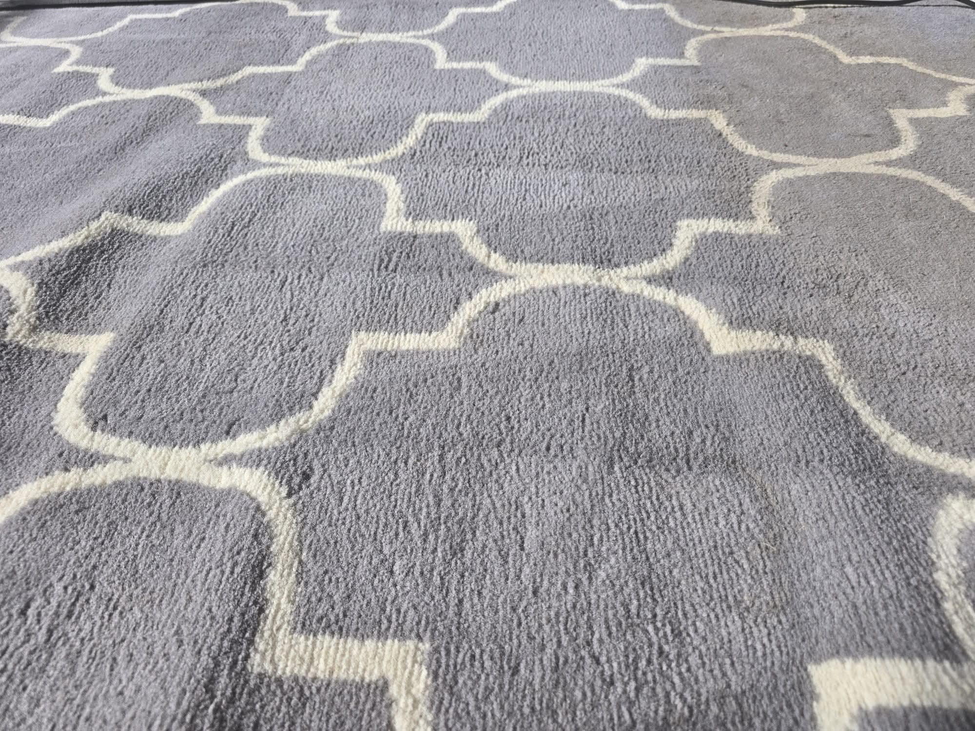 Szary dywan z geometrycznym wzorem w kształcie koniczyny, wykonany z miękkiego, puszystego materiału. Widoczne włókna i struktura dywanu.